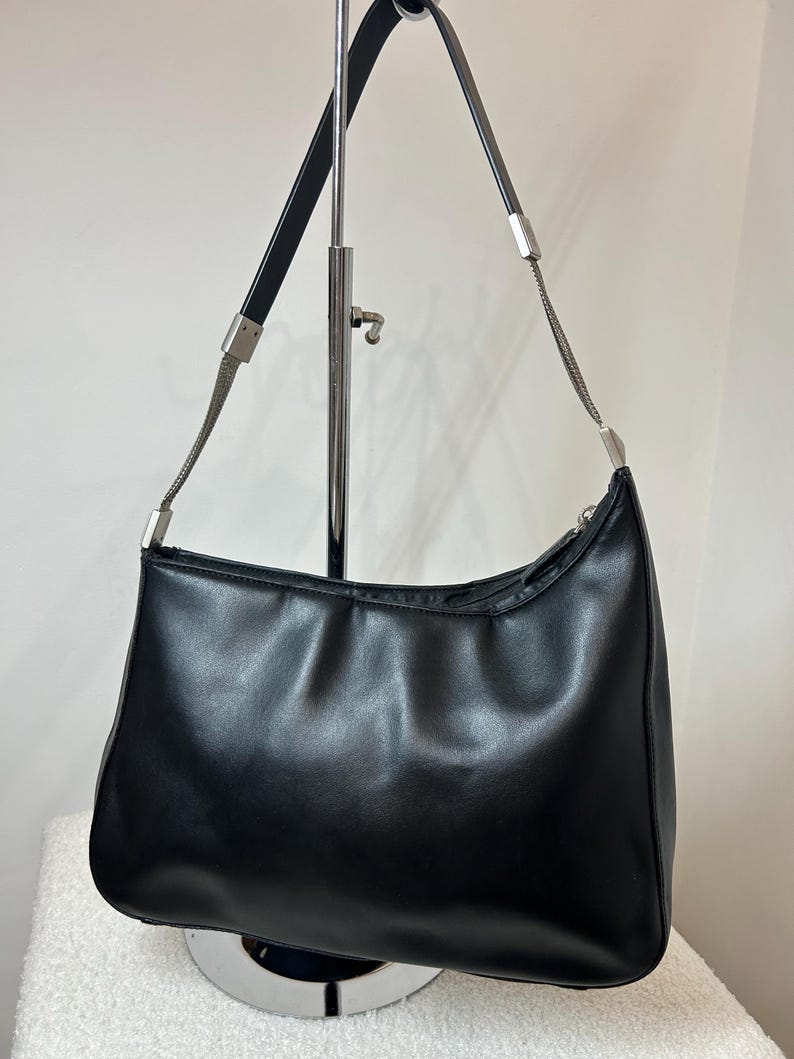 May include: Black shoulder bag with a curved design. The bag features a silver chain and black leather straps. The bag is displayed on a silver stand. The bag has a zipper closure.