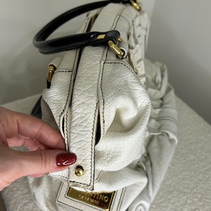 May include: White leather handbag with black stitching and a black handle. The bag features gold-colored hardware and a rectangular gold-colored label with the text "Valentino Garavani". The bag has a textured surface.