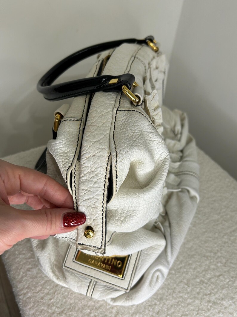 May include: White leather handbag with black stitching and a black handle. The bag features gold-colored hardware and a rectangular gold-colored label with the text "Valentino Garavani". The bag has a textured surface.
