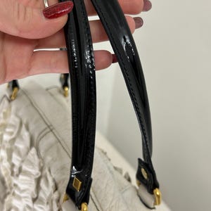 May include: Close-up of a white handbag with black patent leather handles. The bag features gold-toned hardware and a textured leather body. The handles are attached with gold-colored metal squares.