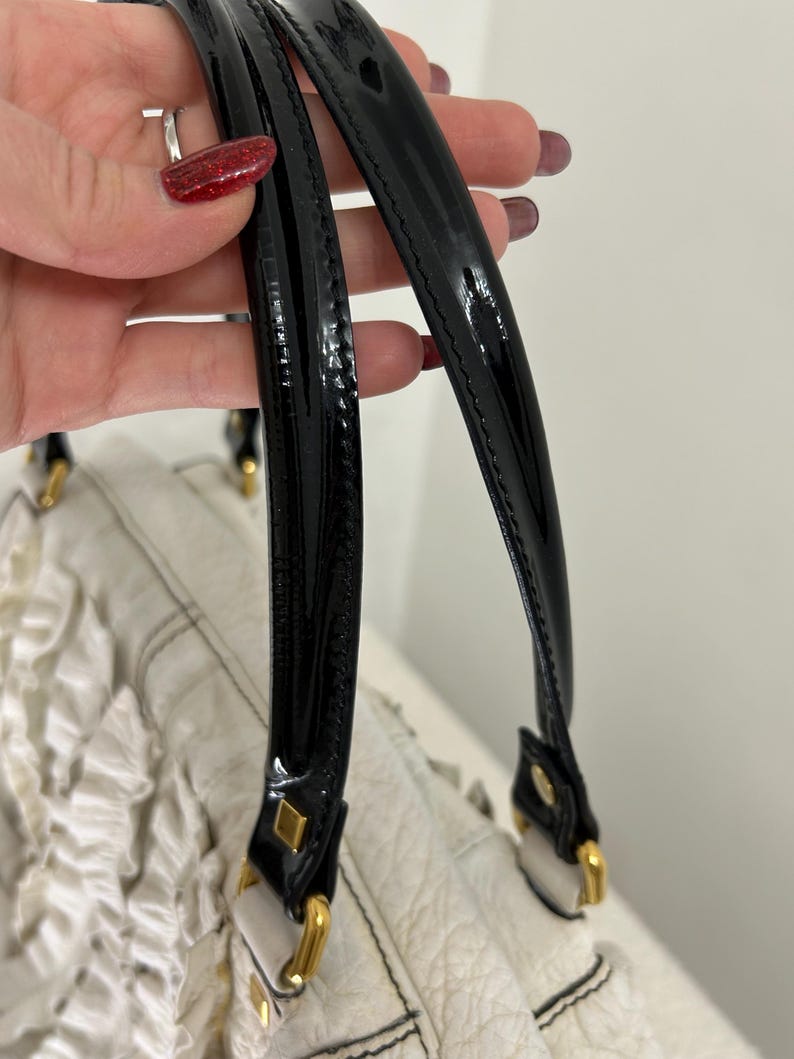 May include: Close-up of a white handbag with black patent leather handles. The bag features gold-toned hardware and a textured leather body. The handles are attached with gold-colored metal squares.