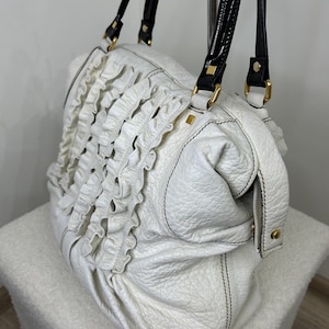 May include: White leather handbag with black handles and gold-tone hardware. The bag features a ruffled design on the front and black stitching. The handles are glossy black with white accents.
