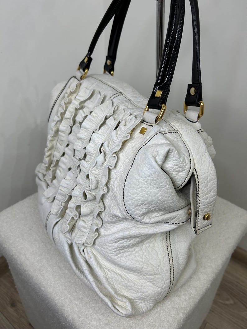 May include: White leather handbag with black handles and gold-tone hardware. The bag features a ruffled design on the front and black stitching. The handles are glossy black with white accents.