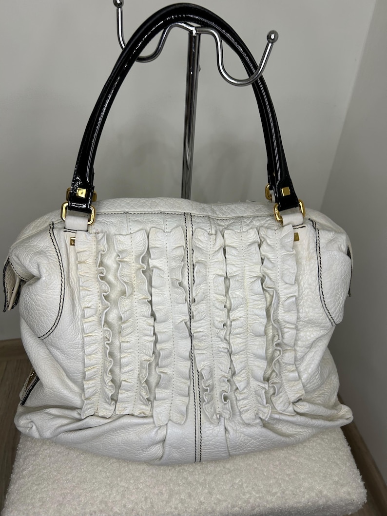 May include: White leather handbag with black stitching and ruffled detailing. Features black patent leather handles with gold-tone hardware. The bag is a soft, slouchy style.