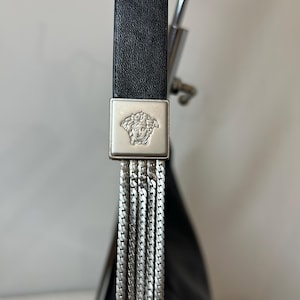 May include: Close-up of a black leather handbag strap with a silver-tone metal square buckle featuring a Medusa head emblem. The strap connects to a series of silver chain tassels, creating a luxurious and stylish design.