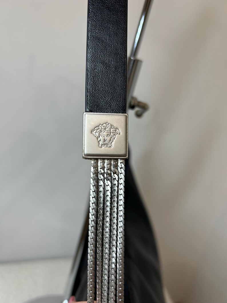 May include: Close-up of a black leather handbag strap with a silver-tone metal square buckle featuring a Medusa head emblem. The strap connects to a series of silver chain tassels, creating a luxurious and stylish design.