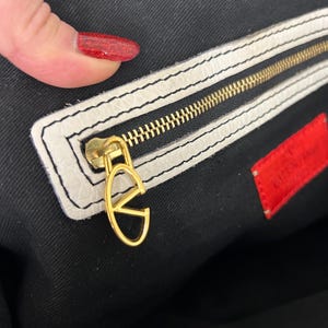 May include: Close-up of a luxury handbag interior. The bag features a gold zipper and a gold 'V' logo charm. The interior is black with a white leather trim. A red tag with white text is visible.