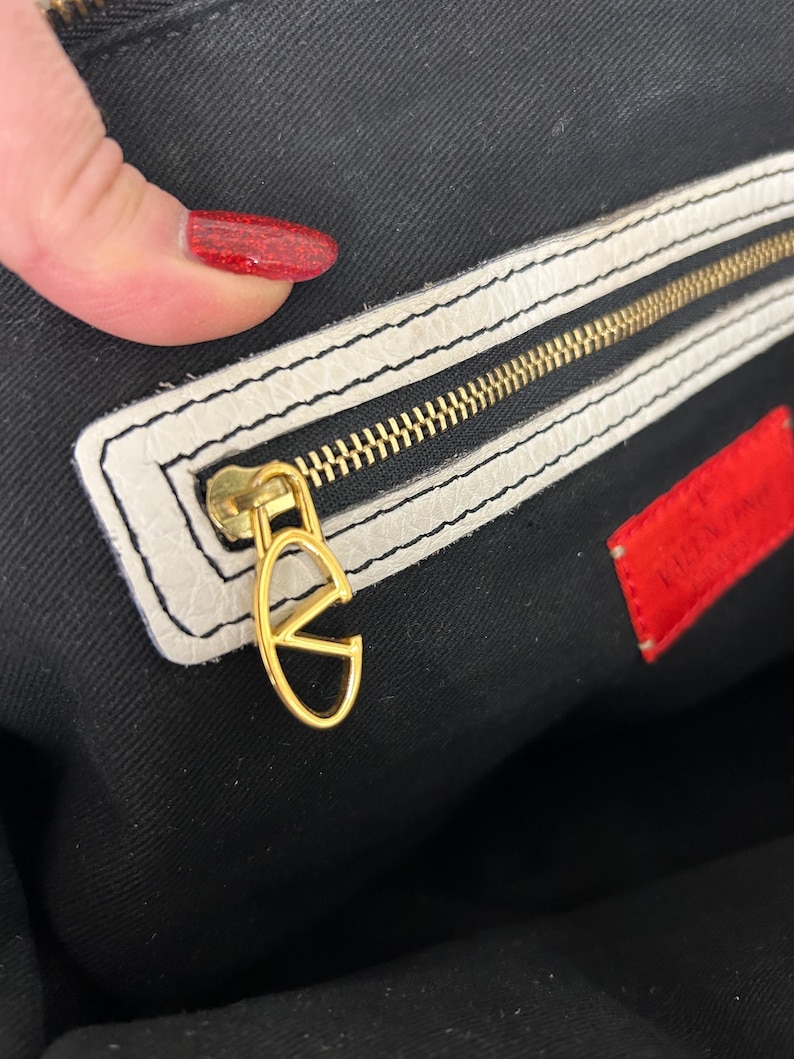 May include: Close-up of a luxury handbag interior. The bag features a gold zipper and a gold 'V' logo charm. The interior is black with a white leather trim. A red tag with white text is visible.