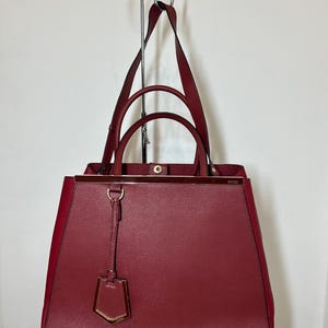 May include: A burgundy leather handbag with a structured design. The bag features a top handle, a detachable tag, and gold-toned hardware. The bag is hanging from a metal hook against a white wall.