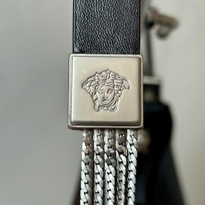 May include: Close-up of a black leather belt with a silver-toned square buckle. The buckle features a Medusa head emblem and a series of dangling silver chain links. The belt is a luxury fashion accessory.