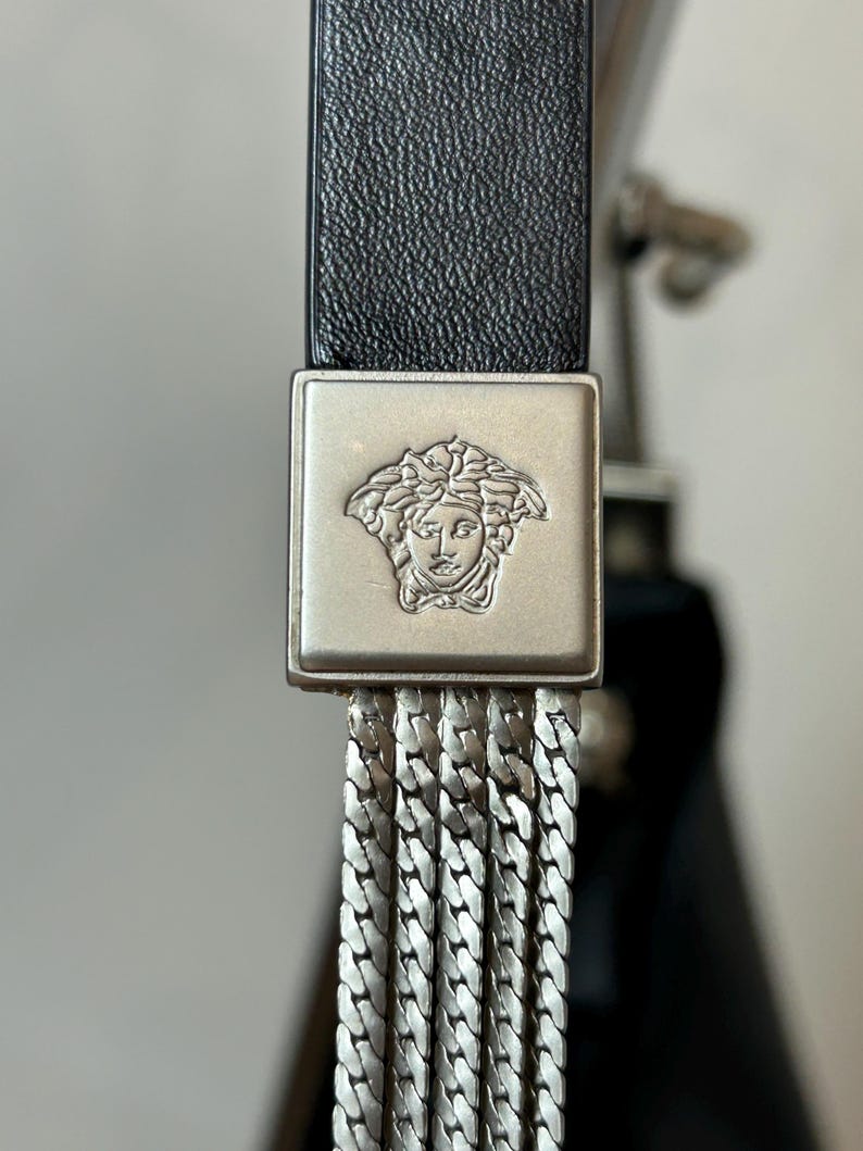 May include: Close-up of a black leather belt with a silver-toned square buckle. The buckle features a Medusa head emblem and a series of dangling silver chain links. The belt is a luxury fashion accessory.