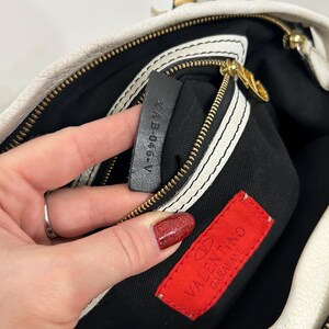May include: Cream-colored handbag with a gold zipper and black interior. The bag features a black leather tag with the text "XAB-046-V" and a red Valentino Garavani label.