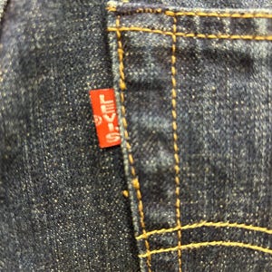 May include: Close-up of a dark blue denim pocket with yellow stitching. A small red tag with the word "LEVI'S" in white lettering is attached to the pocket. The denim has a textured appearance.