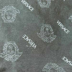 May include: Close-up of a black fabric with a repeating pattern of the Versace logo and Medusa head in white. The fabric has a subtle textured appearance, with diagonal lines adding depth to the design.