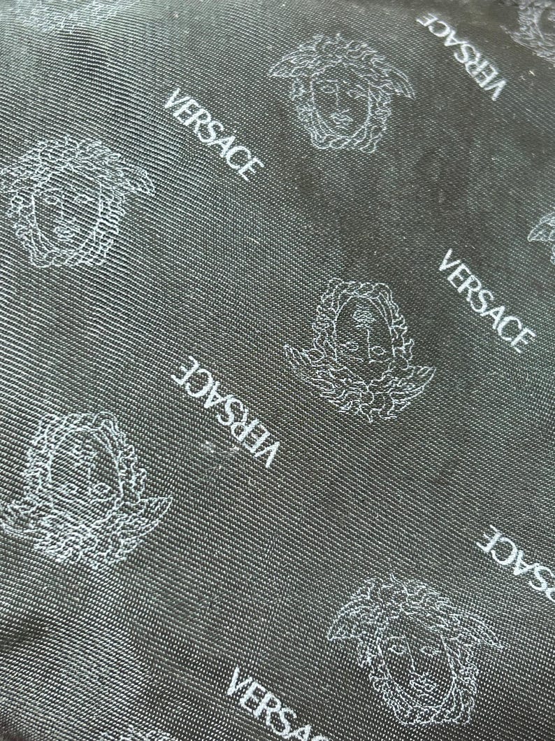 May include: Close-up of a black fabric with a repeating pattern of the Versace logo and Medusa head in white. The fabric has a subtle textured appearance, with diagonal lines adding depth to the design.