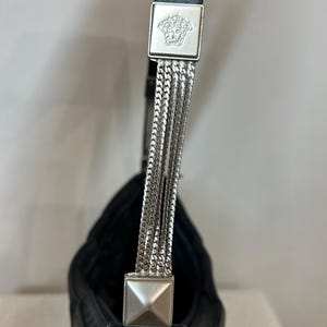 May include: Black leather shoulder bag with silver chain strap detail. The bag features a square silver emblem with a Medusa head logo. The strap is attached with silver chain links.