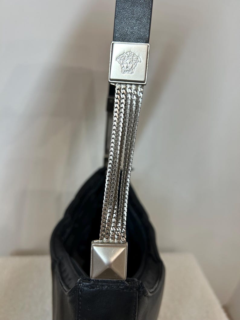 May include: Black leather shoulder bag with silver chain strap detail. The bag features a square silver emblem with a Medusa head logo. The strap is attached with silver chain links.