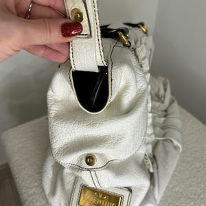 May include: White leather handbag with black stitching and gold-tone hardware. The bag features a top handle, a snap closure, and a gold rectangular plaque with the brand name "Valentino Garavani". The interior is black.