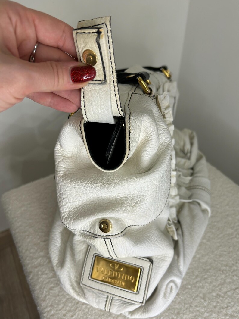 May include: White leather handbag with black stitching and gold-tone hardware. The bag features a top handle, a snap closure, and a gold rectangular plaque with the brand name "Valentino Garavani". The interior is black.