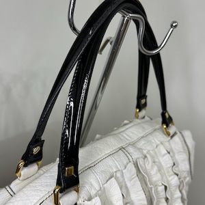 May include: White handbag with black patent leather handles and gold-tone hardware. The bag features a textured body with vertical ruffle detailing. The handles are attached with gold-tone metal clasps. The bag is hanging on a chrome rack.
