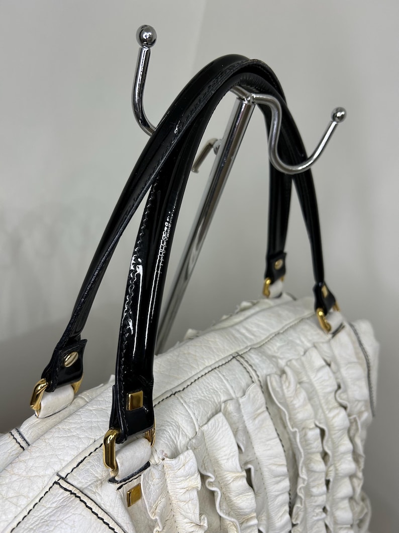 May include: White handbag with black patent leather handles and gold-tone hardware. The bag features a textured body with vertical ruffle detailing. The handles are attached with gold-tone metal clasps. The bag is hanging on a chrome rack.