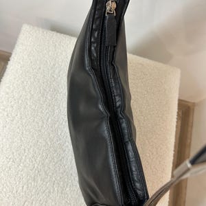 May include: Black leather shoulder bag with a silver zipper and strap hardware. The bag is slightly curved and has a zipper closure. The strap is a light brown color.