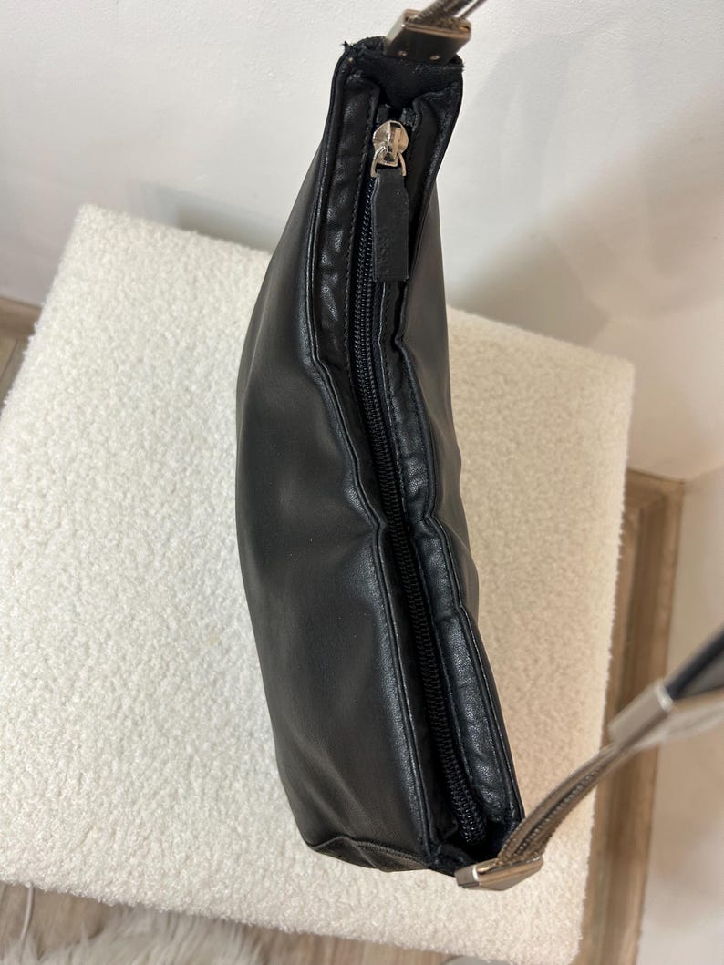 May include: Black leather shoulder bag with a silver zipper and strap hardware. The bag is slightly curved and has a zipper closure. The strap is a light brown color.