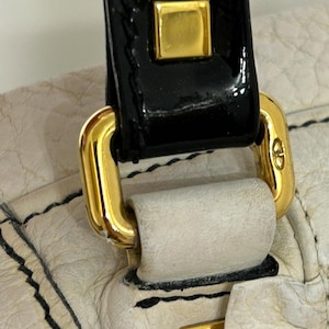 May include: Close-up of a cream-colored leather handbag with black stitching. The bag features a black patent leather handle with gold-colored metal hardware, including a square accent and a clasp.