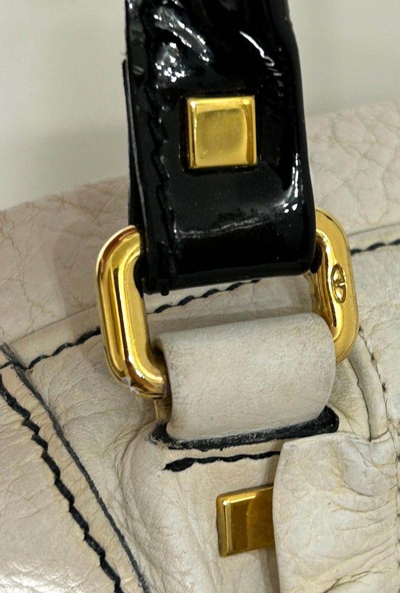May include: Close-up of a cream-colored leather handbag with black stitching. The bag features a black patent leather handle with gold-colored metal hardware, including a square accent and a clasp.