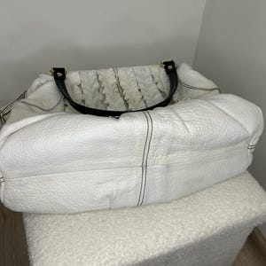 May include: A white leather handbag with black handles and gold-tone hardware. The bag has a textured surface and a gathered fabric detail on the top. Black stitching accents the seams of the bag. The bag is sitting on a white, textured ottoman.