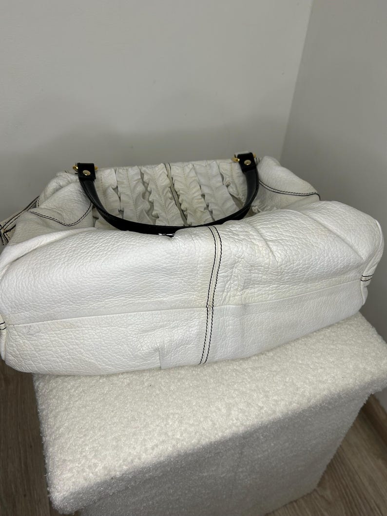 May include: A white leather handbag with black handles and gold-tone hardware. The bag has a textured surface and a gathered fabric detail on the top. Black stitching accents the seams of the bag. The bag is sitting on a white, textured ottoman.