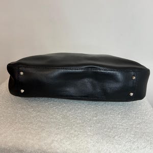 May include: A black leather handbag with a rectangular shape. The bag has a smooth texture and silver-colored studs on the bottom corners. The bag is sitting on a white, textured surface.
