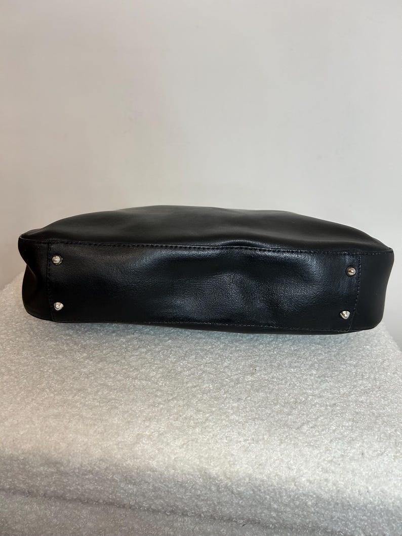 May include: A black leather handbag with a rectangular shape. The bag has a smooth texture and silver-colored studs on the bottom corners. The bag is sitting on a white, textured surface.