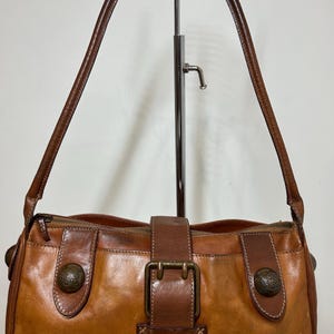 May include: A medium-brown leather shoulder bag featuring a central buckle closure, two side leather tabs with circular metallic decorative studs, a top zipper, and a single shoulder strap. The leather has a distressed, multi-tonal finish.