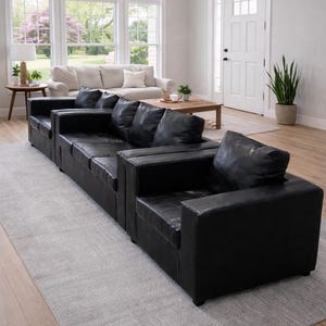 May include: A black leather sectional sofa with several cushions, situated on a light grey rug in a living room. Another sofa is visible in the background, alongside a wooden coffee table and a potted plant. The room has wooden flooring.