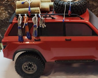 May include: A red toy SUV with a black roof rack carrying wooden logs, a spare tire, and a white bag. Bundles of rope in various colors hang from the rack. The text "theRCscaleRopeShop.etsy.com" is at the bottom.