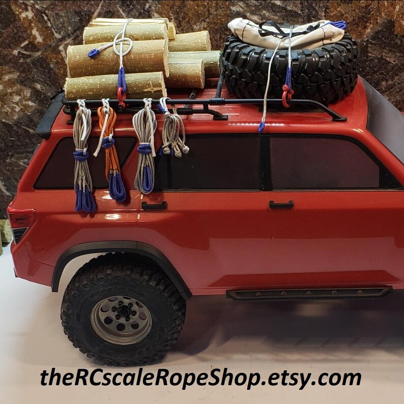 May include: A red toy SUV with a black roof rack carrying wooden logs, a spare tire, and a white bag. Bundles of rope in various colors hang from the rack. The text "theRCscaleRopeShop.etsy.com" is at the bottom.