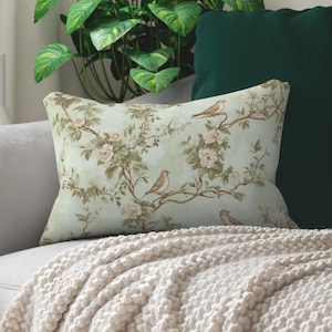 May include: A rectangular decorative pillow with a light green background featuring a floral and bird pattern. The design includes beige birds perched on branches with pink and white flowers. The pillow is placed on a gray sofa with a cream-colored knitted throw.
