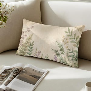 May include: Rectangular beige pillow with a botanical design featuring green and purple leaves and flowers. The pillow is placed on a light-colored sofa, with an open magazine in the foreground.