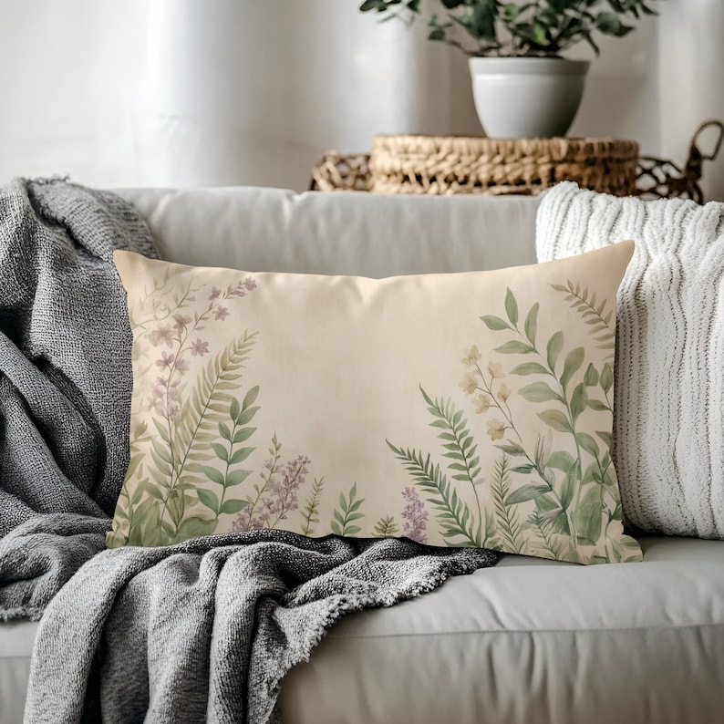 May include: Rectangular decorative pillow with a beige background and watercolor floral design. The pillow features green ferns, lavender, and white flowers. The pillow is on a gray sofa with a gray blanket.