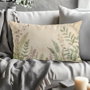 May include: Rectangular decorative pillow with a beige background and a watercolor floral design. The design features green ferns, purple flowers, and white blossoms. The pillow is placed on a gray sofa with a gray blanket.