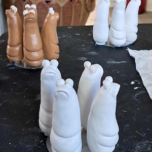 May include: A collection of ceramic sculptures in various stages of completion. Some are a light brown color, while others are white. The sculptures have a rounded, abstract form with a small, rounded top. They are arranged on a black surface.