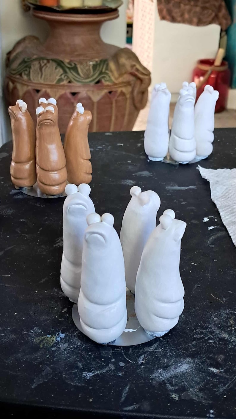May include: A collection of ceramic sculptures in various stages of completion. Some are a light brown color, while others are white. The sculptures have a rounded, abstract form with a small, rounded top. They are arranged on a black surface.