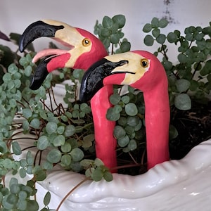 May include: Two pink flamingo head garden stakes with black and cream beaks, one with an open mouth showing a tongue, positioned among green foliage in a white textured planter. These decorative figures feature realistic eyes and smooth surfaces.