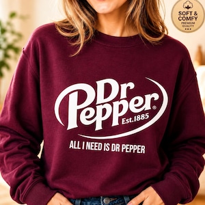 May include: A maroon long-sleeve sweatshirt featuring the white Dr Pepper logo with the text Est.1885 and the phrase ALL I NEED IS DR PEPPER printed below. A circular badge in the top right corner contains the text SOFT & COMFY PREMIUM QUALITY.