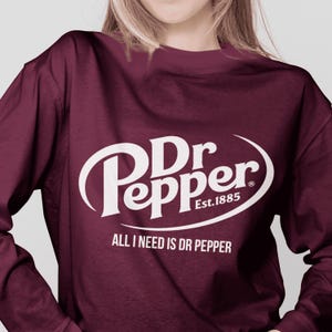 May include: A maroon long-sleeve shirt featuring the Dr Pepper logo in white. The logo includes the text "Dr Pepper Est.1885" and the phrase "ALL I NEED IS DR PEPPER" in white lettering.