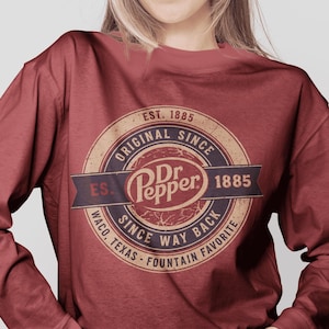 May include: A maroon long-sleeve shirt featuring a circular graphic with the text "Dr Pepper" in a white oval. The graphic includes the words "Original Since," "Est. 1885," and "Waco, Texas - Fountain Favorite."