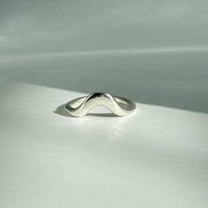 Puede incluir: Un anillo plateado con un diseño ondulado. El anillo tiene una superficie lisa y pulida y una forma curva, que recuerda a una ola estilizada. El anillo está sobre un fondo blanco, con sombras que añaden profundidad.