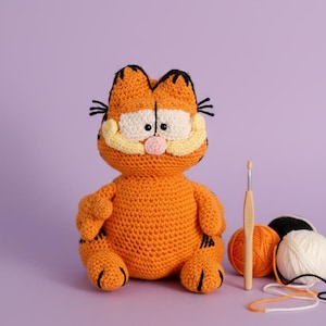 Puede incluir: Peluche de Garfield naranja tejido a ganchillo, con detalles negros, sobre un fondo morado. El peluche tiene ojos blancos, hocico amarillo y blanco, y nariz rosa. Se ven también ovillos de lana y un ganchillo.
