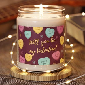 Puede incluir: Una vela encendida en un tarro de cristal con la frase "¿Quieres ser mi San Valentín?" y caramelos en forma de corazón. La vela está sobre un pequeño posavasos de madera, con luces de cadena.